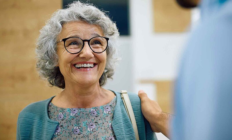 Eine ältere Frau lächelt und blickt zu einer weiteren Person, die ihr eine hand auf die Schulter legt.